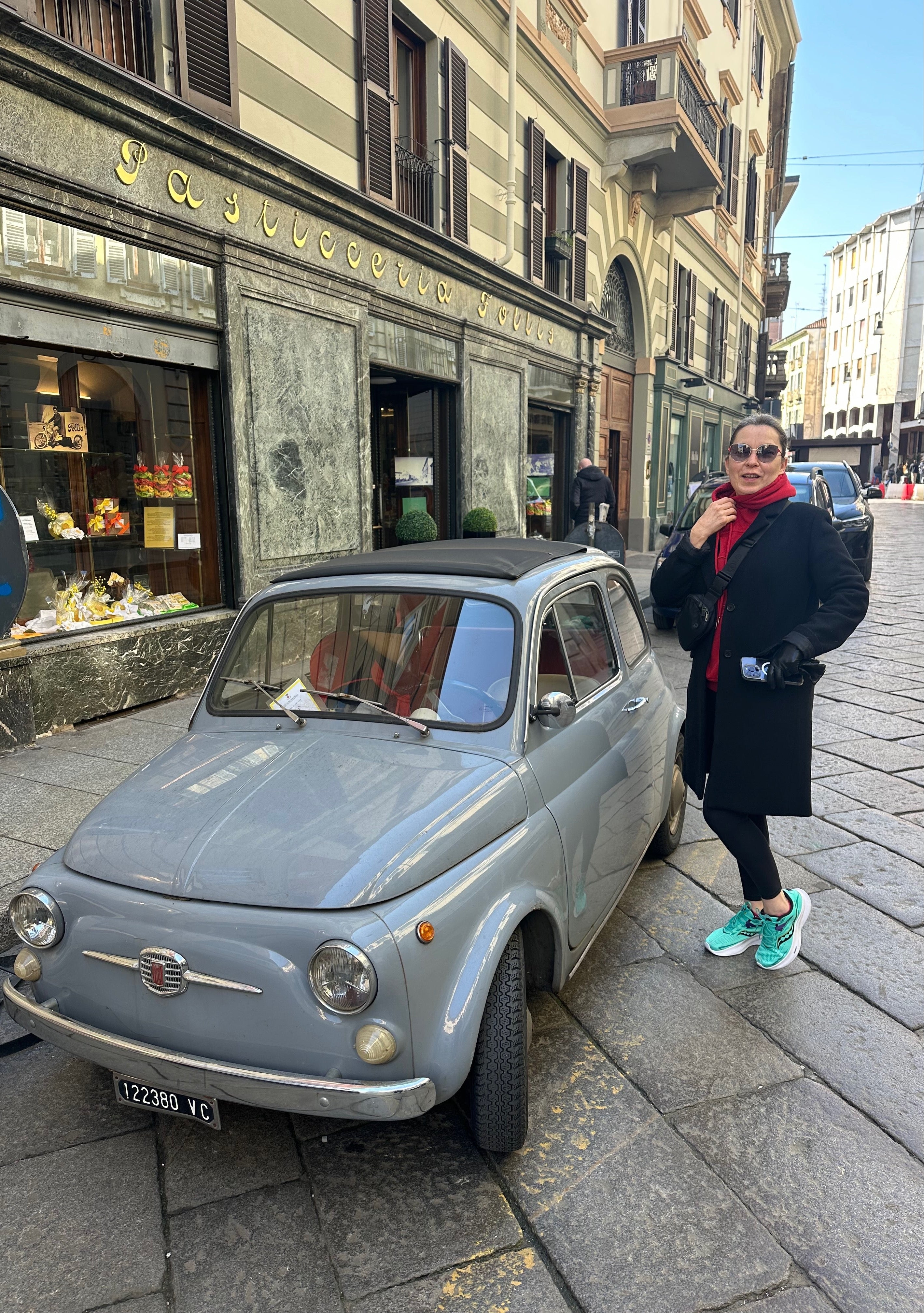 A woman named Oxana standing next to an old Fiat car on a street in Italy.